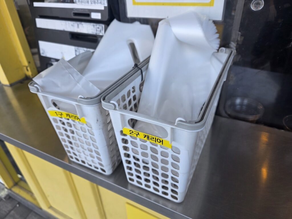 5 Unique Rituals of Mega Coffee: Beyond the K-Pop Idol Diet showing white plastic baskets labeled for 1-cup and 2-cup vinyl carriers at a pickup counter.