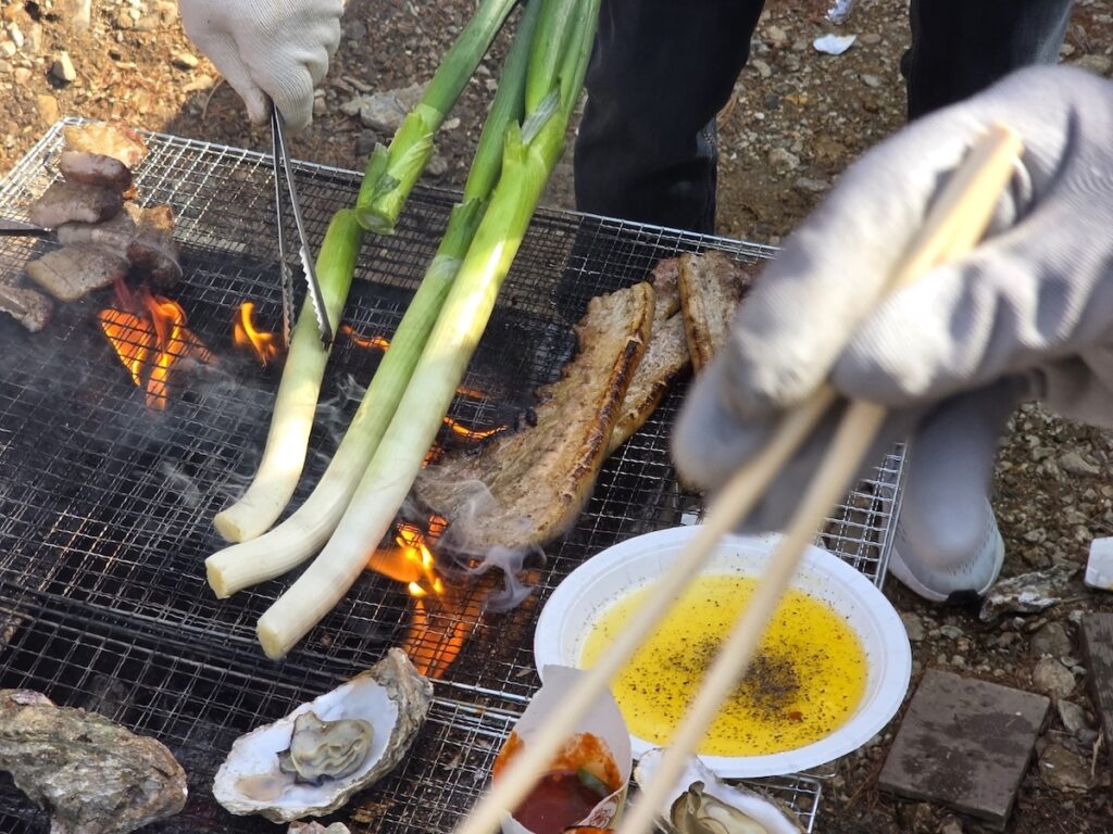 Authentic Perilla Oil Sauce sits ready in a white bowl next to pork, scallions, and oysters roasting on an open-fire grill.