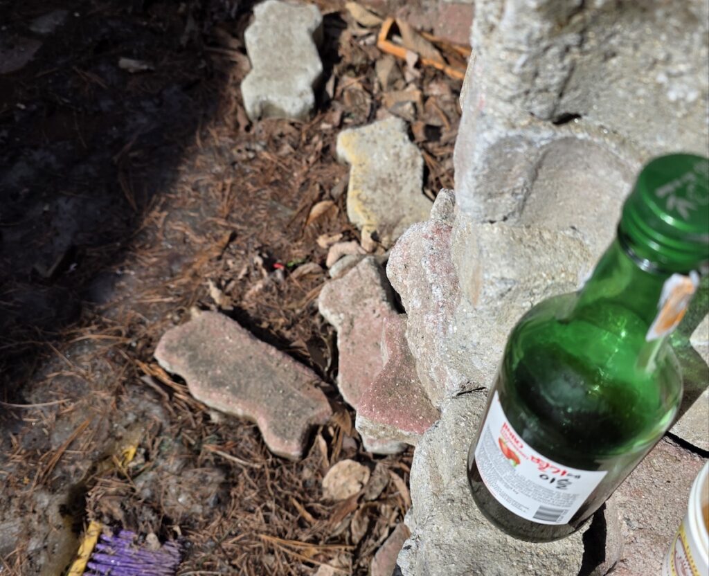 A recycled green soju bottle filled with dark, toasted Authentic Perilla Oil Sauce resting on a rustic stone wall.