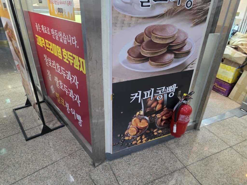 The entrance of a 10 Trendy Coffee Bean Bread Rituals in Gangnam stall located right outside a Gangnam subway station exit in Seoul, South Korea. The photo shows the warm lighting, a sign in Korean advertising Keopi-kong-ppang and Hodu-gwaja (walnut cakes), and a red fire extinguisher on the wall.