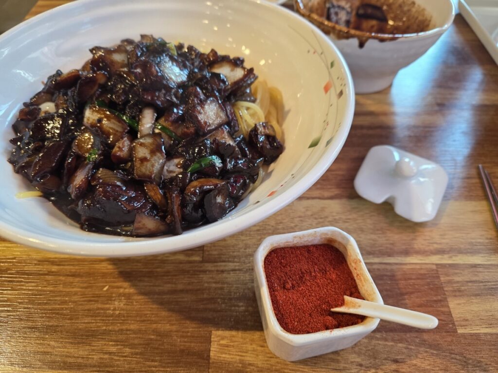 A cinematic shot of Ganjajang, authentic Korean black bean noodles, with a side of vibrant red Gochugaru chili powder.