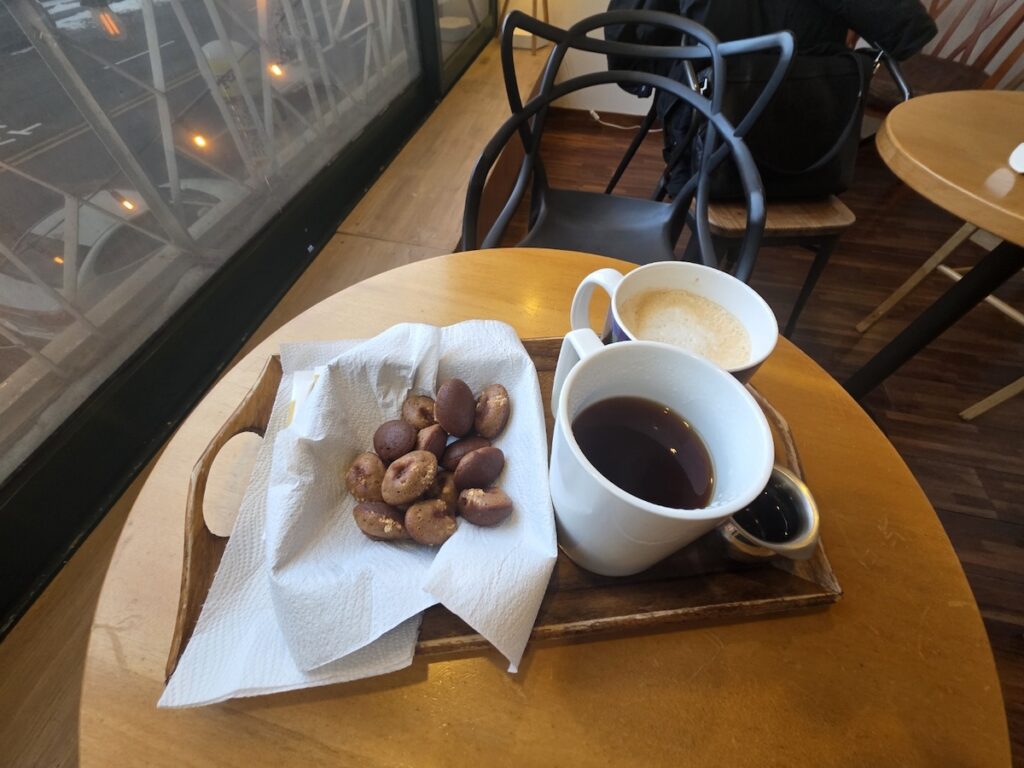 A perspective shot from a cafe window table overlooking a Seoul street at twilight, showing a wooden tray with a basket of Keopi-kong-ppang (Korean coffee bean bread) and two ceramic mugs—one filled with hot Black Coffee (Long Black) and the other with a Caffe Latte. This setup embodies one of the 10 Trendy Coffee Bean Bread Rituals in Gangnam, demonstrating the superior pairing with Black Coffee over a Latte.