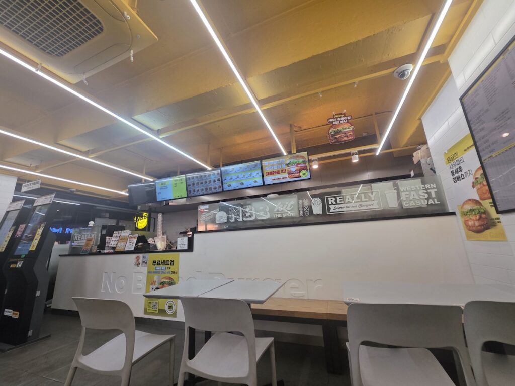 The clean and modern interior of a No Brand Burger (NBB) shop, showing the order counter and high-tech menu boards as part of the Seoul Gourmet Guide 2026.