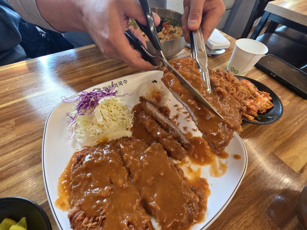 Two hands dynamically using heavy-duty kitchen scissors and tongs to perform surgical-like cuts on a steaming King-sized Donkatsu, illustrating the practical Korean Scissors Culture in action.