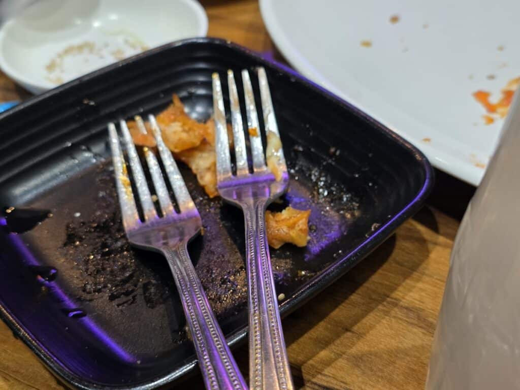 The satisfying aftermath of a Korean chicken meal, showing two used silver forks resting on a black plate after a successful dual-fork bone removal ritual.