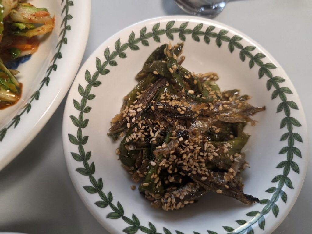 A top-down view of a small side dish of stir-fried anchovies with green chilis (Myeolchi-gochu-bokkeum). This mineral-rich Korean weight loss diet banchan is generously coated in a sweet-savory glaze and covered in sesame seeds, presented in a signature patterned bowl.