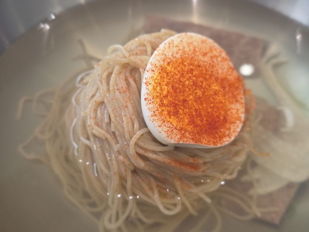 A close-up cinematic shot focusing on the coiled ball of hand-pressed buckwheat noodles in a bowl of Pyeongyang naengmyeon. The noodles form a tight, structured mass that resembles red velvet, symbolizing the cultural and political diplomatic story connecting Red Velvet (K-pop) to Okryu-gwan. The surrounding clear broth represents the concentrated Korean meat broth secrets.