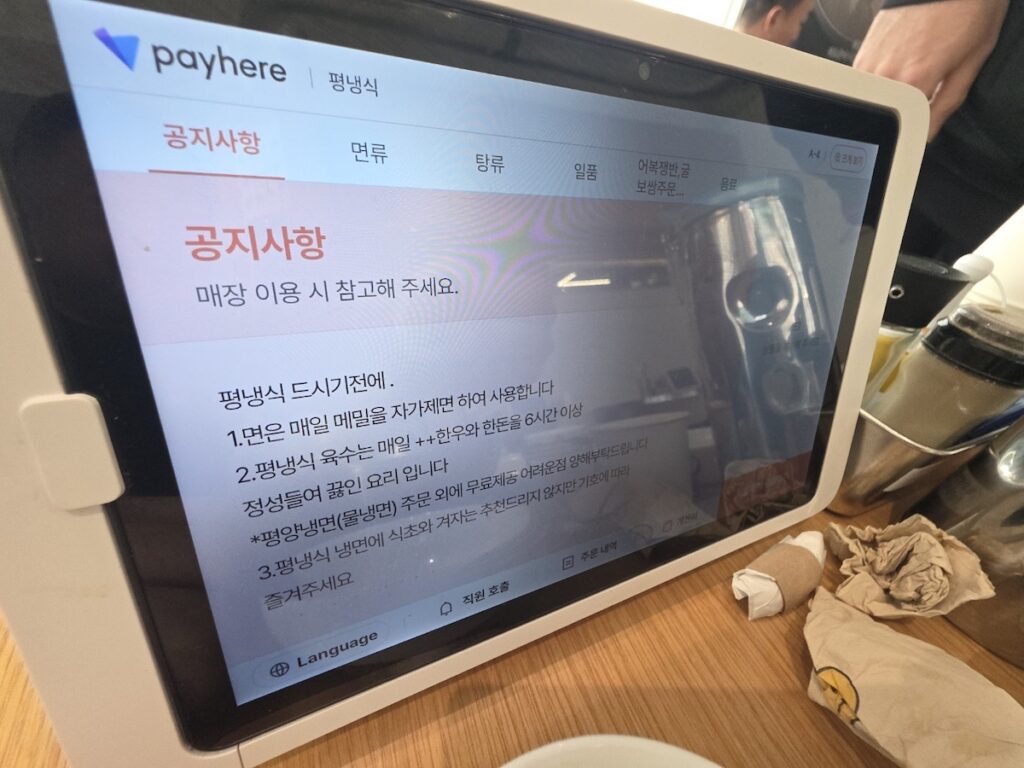 An angled photograph of a digital tablet menu board displaying notice (공지사항) in Korean. The text confirms the use of high-quality "1++ Hanwoo beef and pork for 6 hours" to create the broth. This image provides authentic proof of the concentrated Korean meat broth secrets discussed in the Pyeongyang naengmyeon guide.