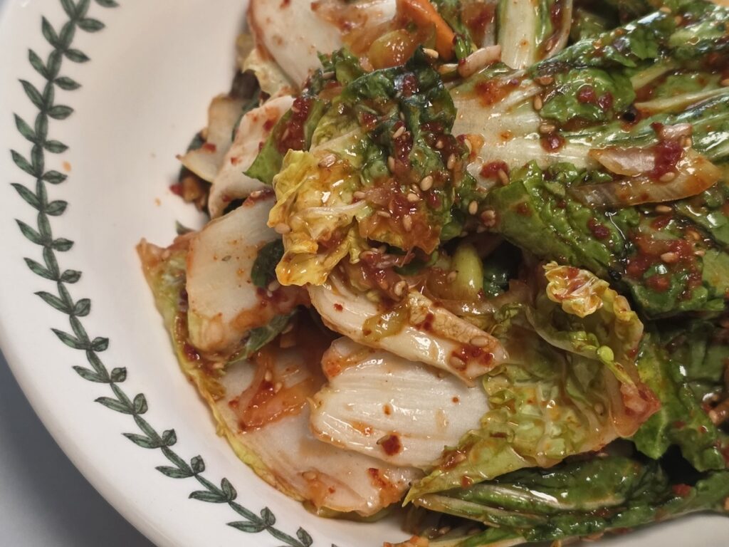 A close-up cinematic shot of Bomdong Geotjeori, a fresh Korean weight loss diet banchan made from early spring cabbage. The vibrant green leaves are lightly tossed in a fiery red chili sauce and sprinkled with toasted sesame seeds, served in a Portmeirion Botanic Garden bowl.