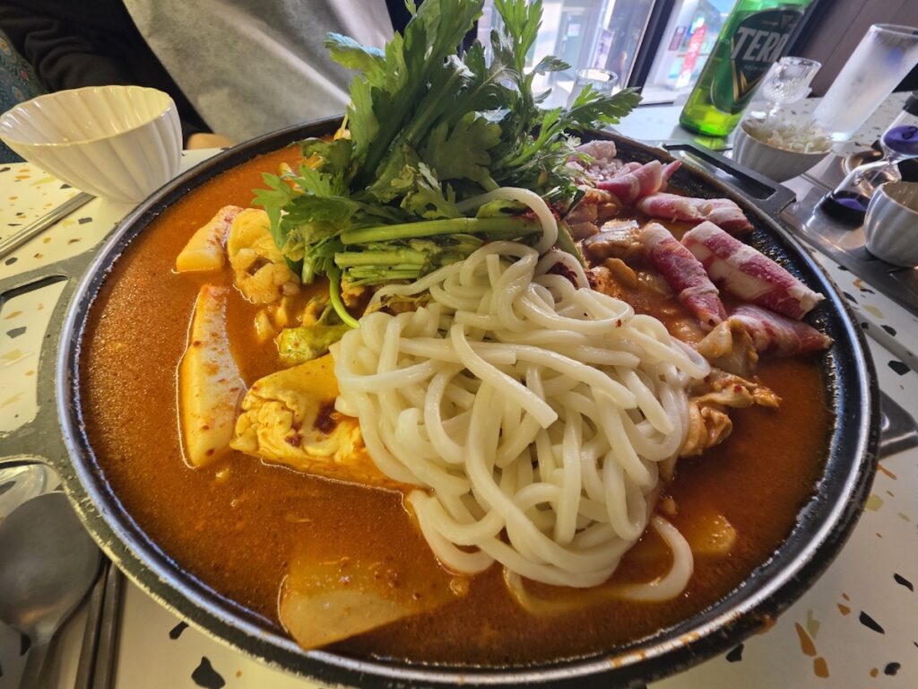 A top-down photograph of a large, black stoneware pot (ttukbaegi) filled with Jjigae (Korean Stew), placed in the center of the table. The pot features udon noodles, beef shabu, large tofu blocks, and is topped with dense mugwort greens. It represents the communal Sharing Food center of the meal, often shared by multiple spoons, contrasting with the individual Guk. A Tera beer bottle is visible in the background.