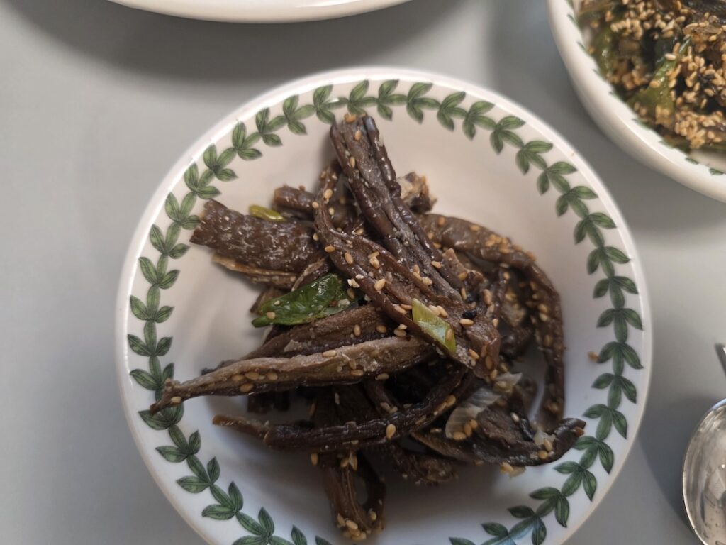 A detailed close-up of dried eggplant stir-fry (Gaji-namul), a satisfying and sophisticated Korean weight loss diet banchan. The dark, chewy texture of the dried eggplant is complemented by slices of green chili and a dusting of sesame seeds in a Portmeirion patterned dish.