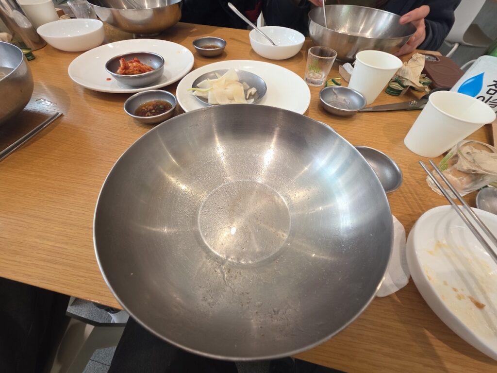 An angled overhead shot of an empty, large stainless steel bowl on a wooden restaurant table in Seoul. The bowl is completely licked clean, surrounded by used side dish plates (Kimchi, radish), a Soju glass, and a metal spoon. This image represents the satisfying completion of the Pyeongyang naengmyeon ritual and the concept of an empty bowl achievement, fulfilling the cultural wisdom discussed in the Seoul healthy food guide.