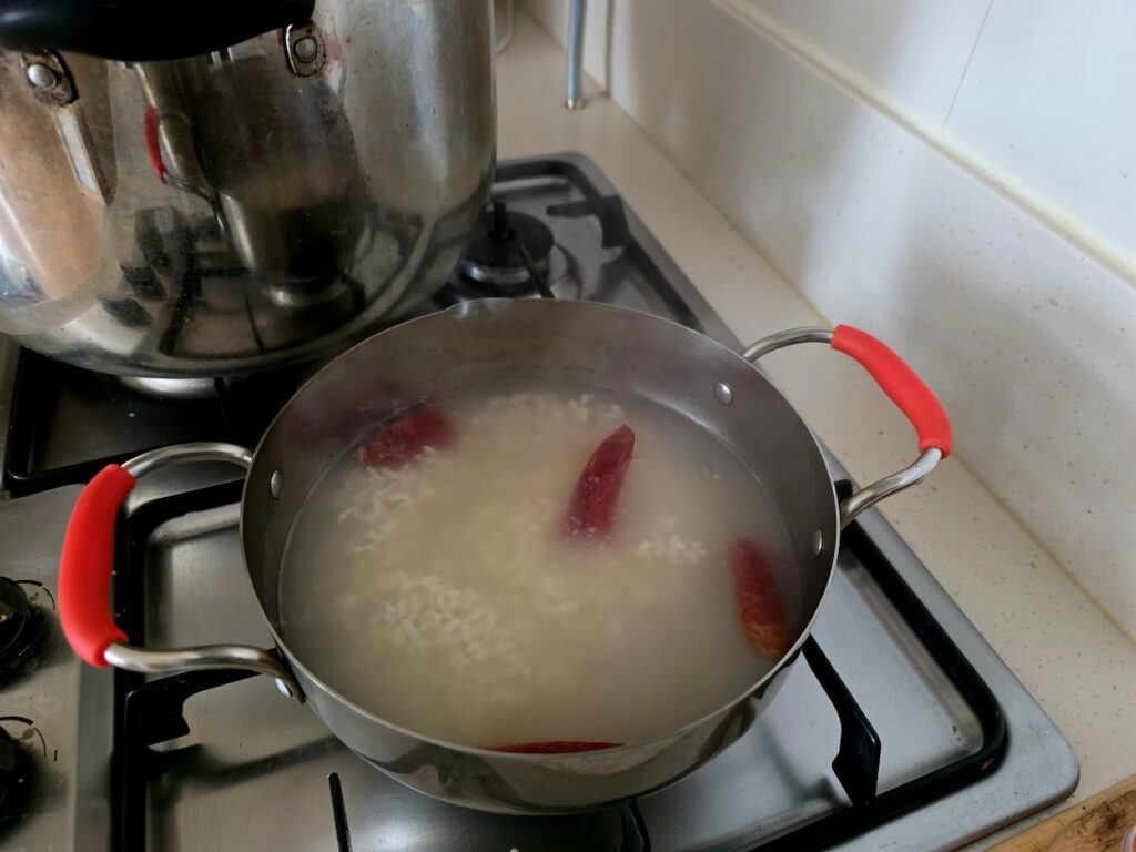 Best Korean Desserts: A stainless steel pot over a gas stove showing the home-brewing process of Sikhye, with multiple dried red chili peppers floating, illustrating the unique spicy family recipe.