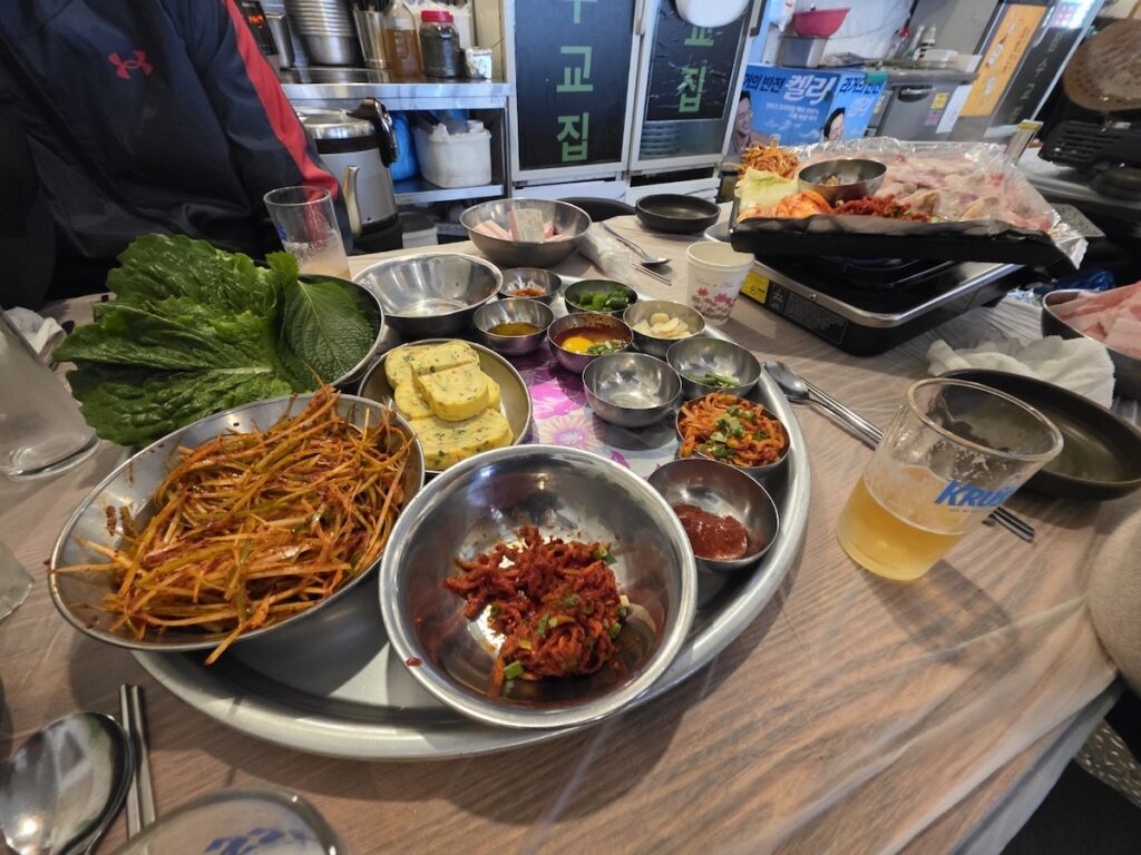 A full table setup of K-BBQ rituals in Seoul featuring various side dishes, fresh lettuce, and pork belly on a burner.