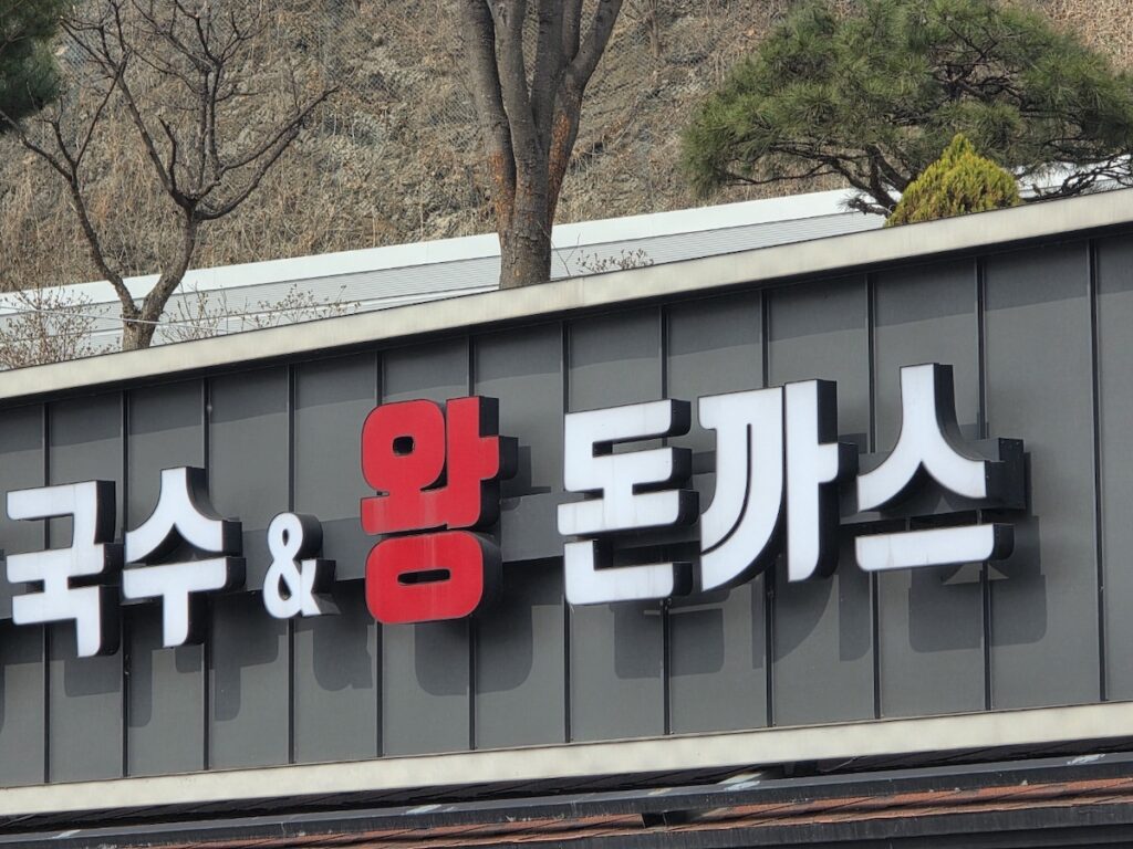 A close-up of a dark grey electronic signboard in Paju Heyri Art Valley, clearly displaying the Korean text for "Wang-Donkatsu" (King-sized Donkatsu) and "Guksu" (Noodles) in white and red.