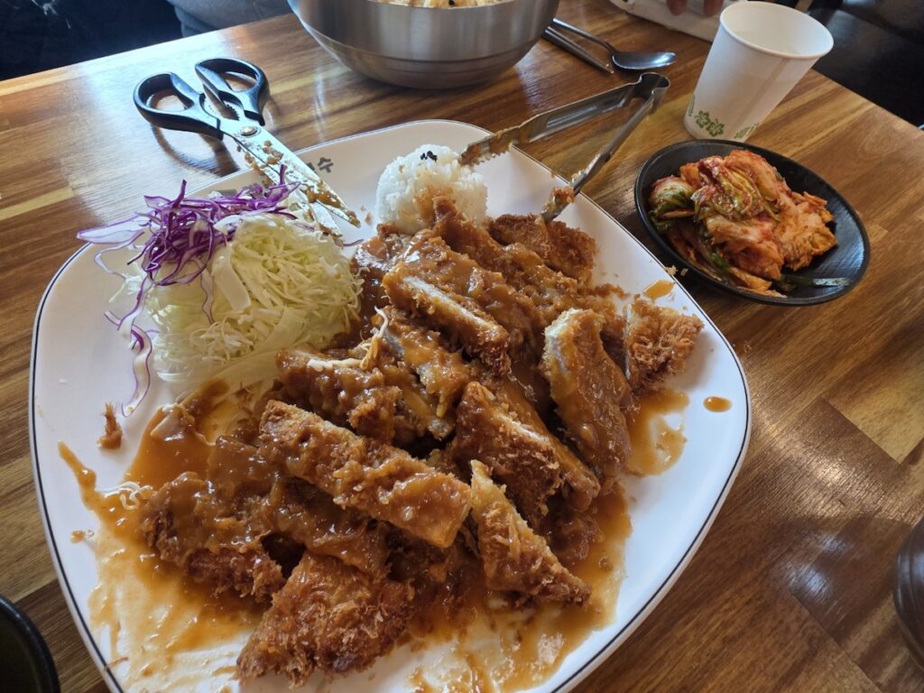 A view of the King-sized Donkatsu after the unique Korean Scissors Ritual is complete, with the massive cutlet processed into bite-sized squares using kitchen scissors and tongs, ready for a communal meal.