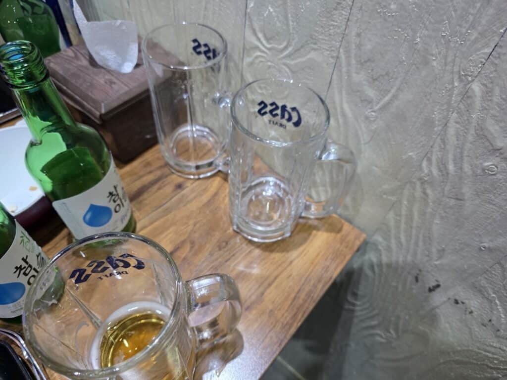 A table setup in a Seoul chicken shop featuring green Soju bottles and multiple 500cc draft mugs, reflecting the collective 'Gun-bae' culture.