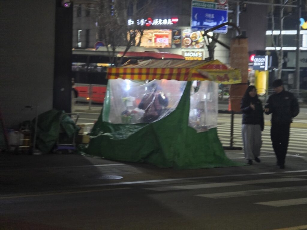 A green and yellow striped street food stall glowing under night lights, offering a secret ritual of Friday night comfort in the Korea Street Food Guide 2026.