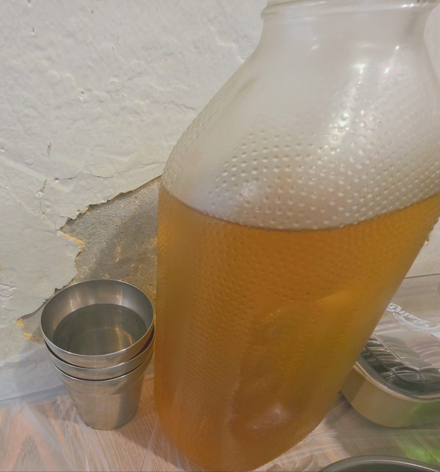 Traditional Korean dining table featuring a vintage Del Monte bottle of barley tea alongside a green soju bottle and stainless steel cups against a rustic, weathered wall, depicting the Social Contract of Korean Barley Tea Culture 2026.
