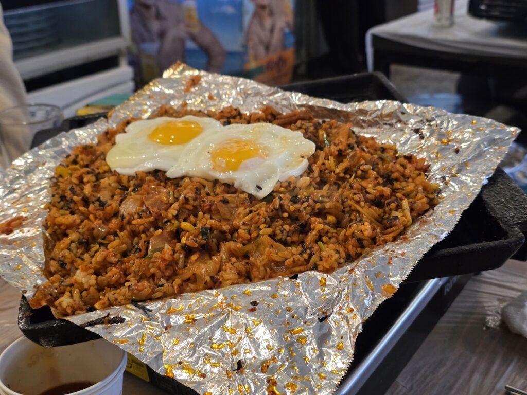 Korean BBQ Fried Rice Ritual: A close-up of a steaming Bokkeumbap topped with two sunny-side-up eggs on a foil-covered grill, illustrating the luxurious finale of a meat-based meal.