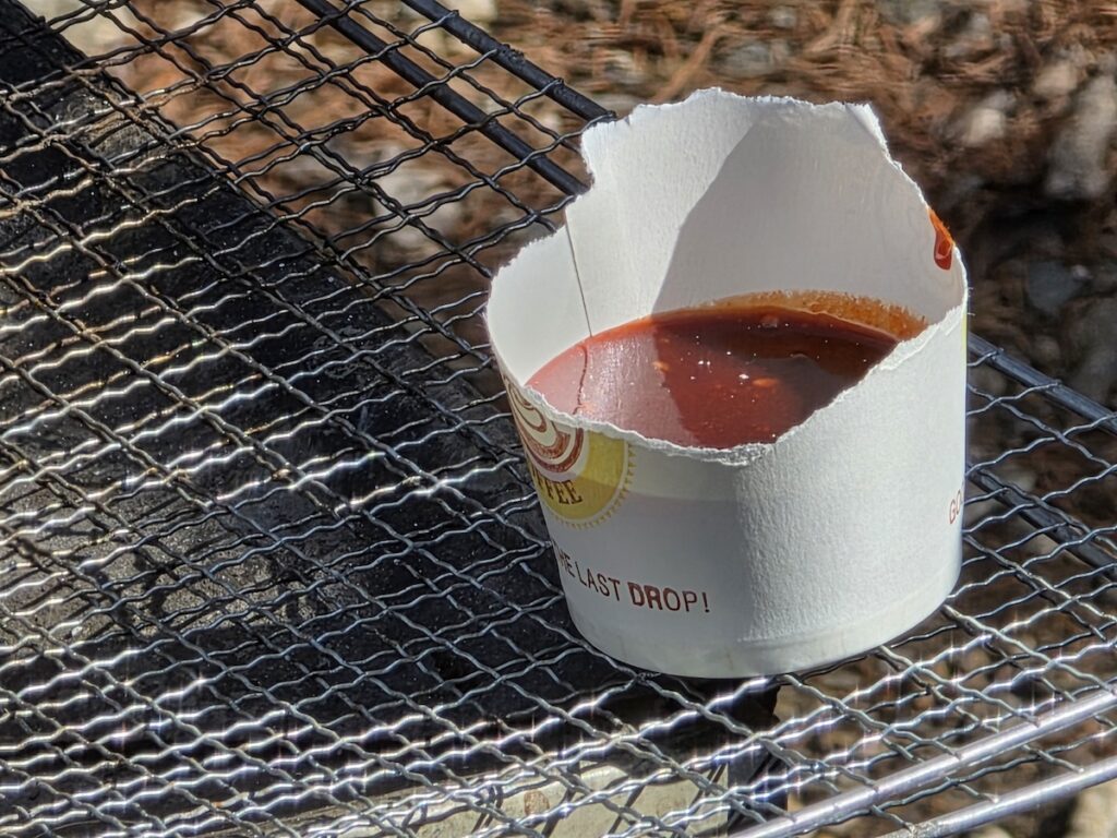 A torn paper cup filled with glossy red Chojang sauce resting on a metal grill, showcasing the rustic street-style ritual within the Korean Chojang Sauce Guide 2026.
