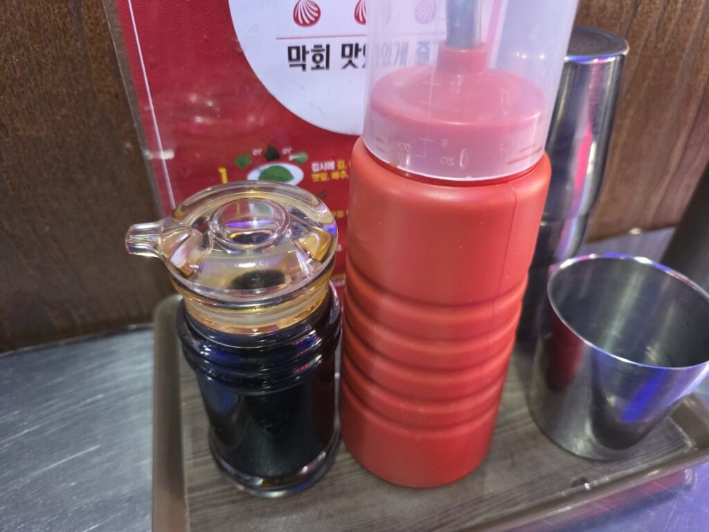 A vibrant red squeeze bottle of Chojang and a soy sauce dispenser on a restaurant table, illustrating the essential condiments in the Korean Chojang Sauce Guide 2026.