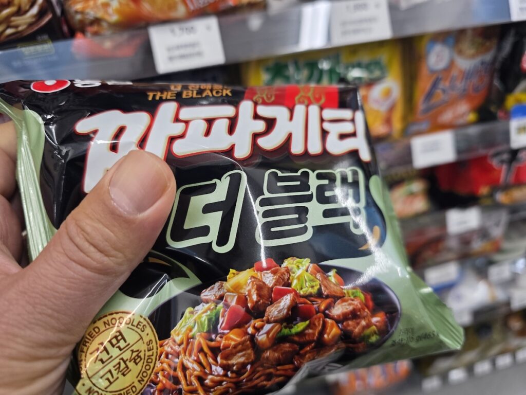 A person holding a pack of Chapagetti at a convenience store, representing the Sunday noodle ritual in Korea.