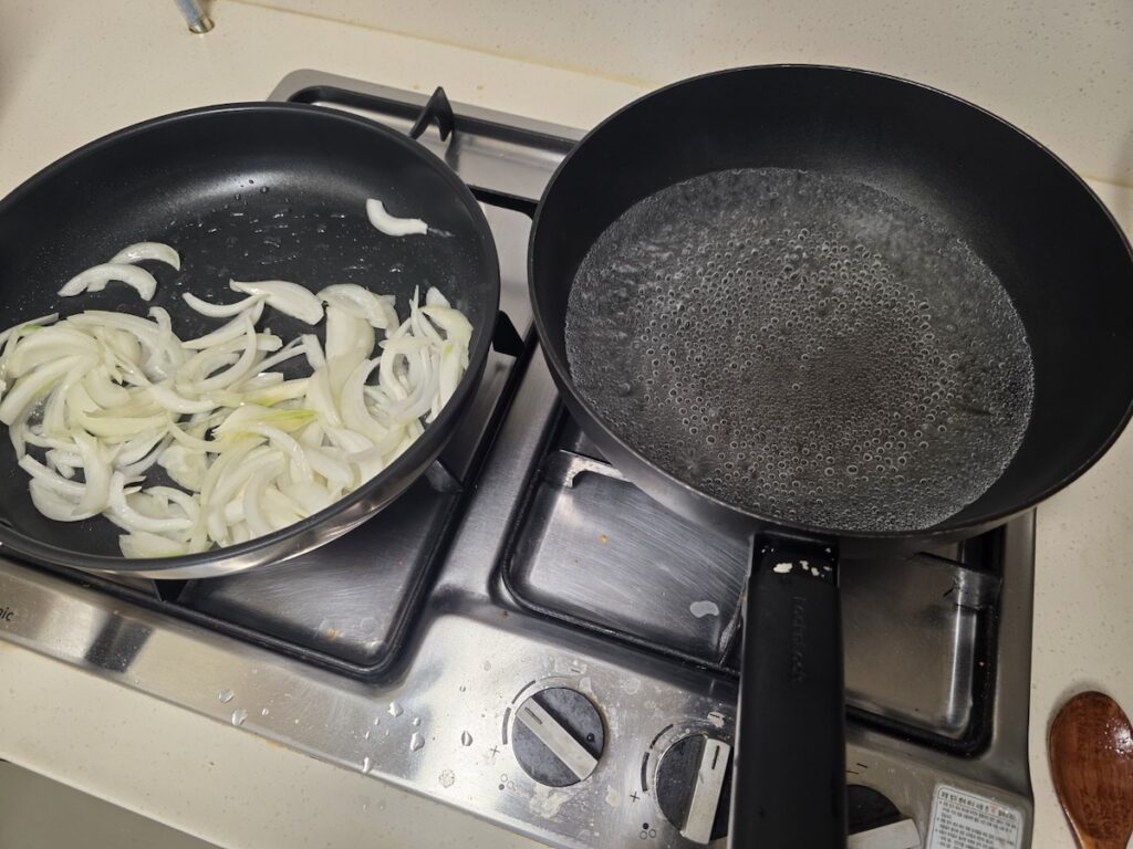 Stir-frying sliced onions on a stove to add depth to the instant noodle ritual of Korean Food Rituals 2026.
