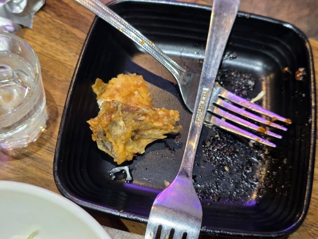 A close-up shot of a crispy piece of Korean fried chicken on a black plate, being dissected by two silver forks to avoid touching the sauce with hands.