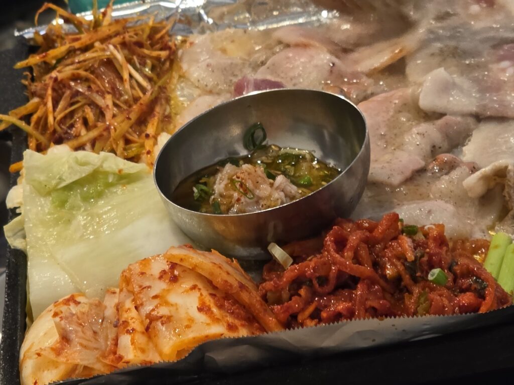 Close-up of frozen pork belly, kimchi, and seasoned scallions sizzling on a slanted griddle in Seoul.
