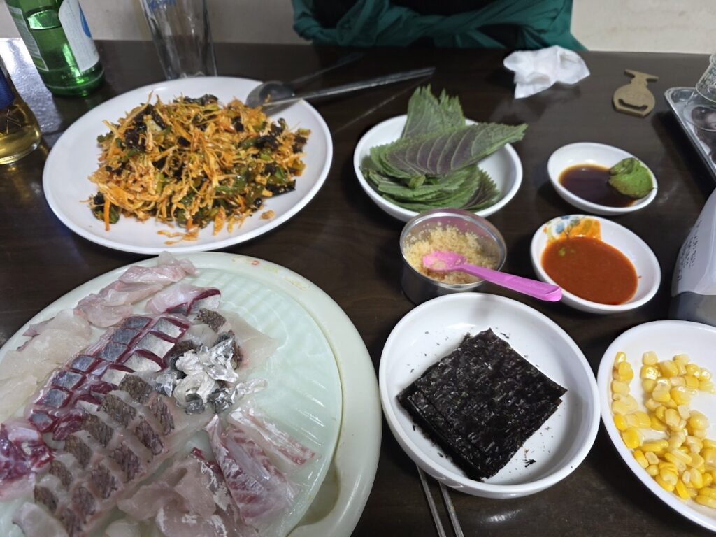 Korean Raw Fish Ssam Ritual: A wide shot of a traditional Seoul raw fish restaurant table, showcasing the harmonious spread of raw fish, perilla leaves, seaweed (Gim), dynamic sauces, and side dishes (banchan) ready for the custom ritual.
