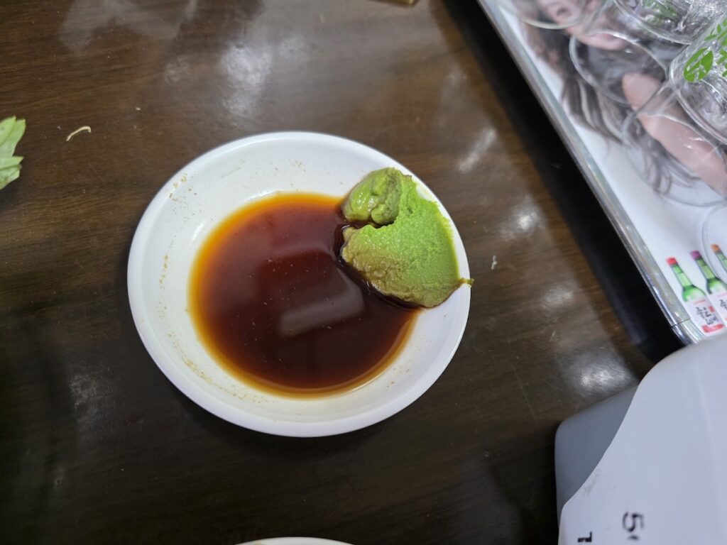 Korean Raw Fish Ssam Ritual: A small dish of dark soy sauce paired with a generous portion of pungent green wasabi, representing the essential starting point of the Korean dipping sequence.