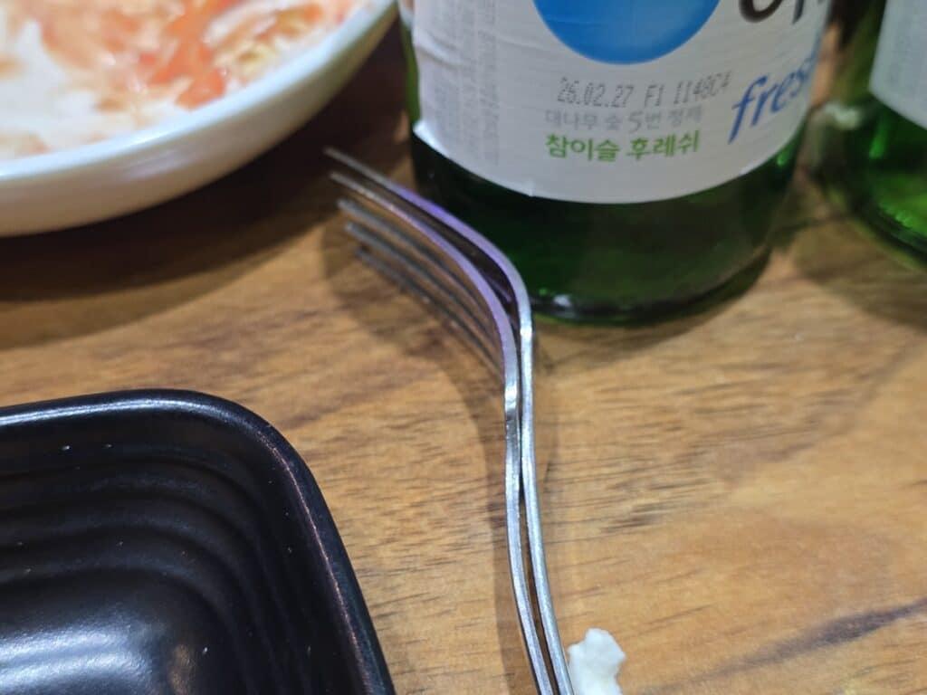 A detailed shot of two silver forks leaning against a green Korean Soju bottle, symbolizing the end of a sophisticated local dining ritual.