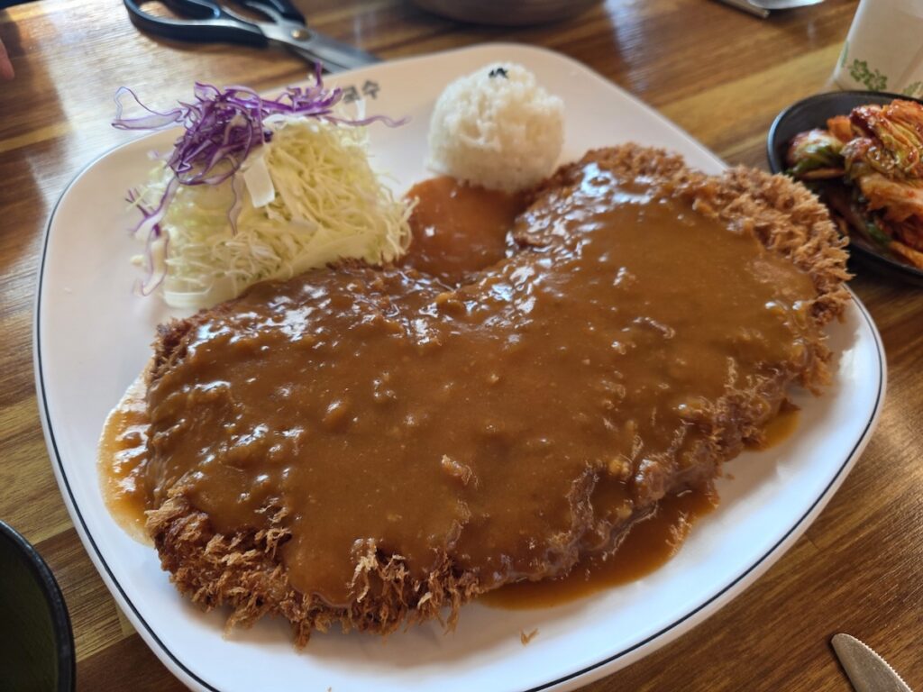 A monstrously large King-sized Donkatsu plated with a rich demi-glace brown sauce, served with a side of cabbage salad and rice, showcasing the ultimate Best Comfort Food in Korea at a Heyri restaurant.