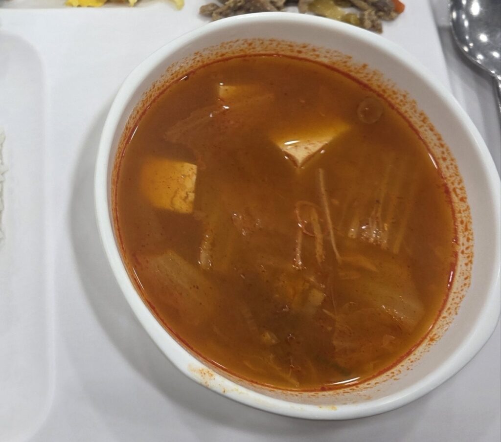A close-up photograph of a private, oval-shaped white bowl filled with Guk (Kimchi Soup), showing delicate tofu cubes and shredded cabbage in a clear broth. This is an individual serving, private to the diner, as referenced in the text comparing Guk vs Jjigae in Korean food culture.