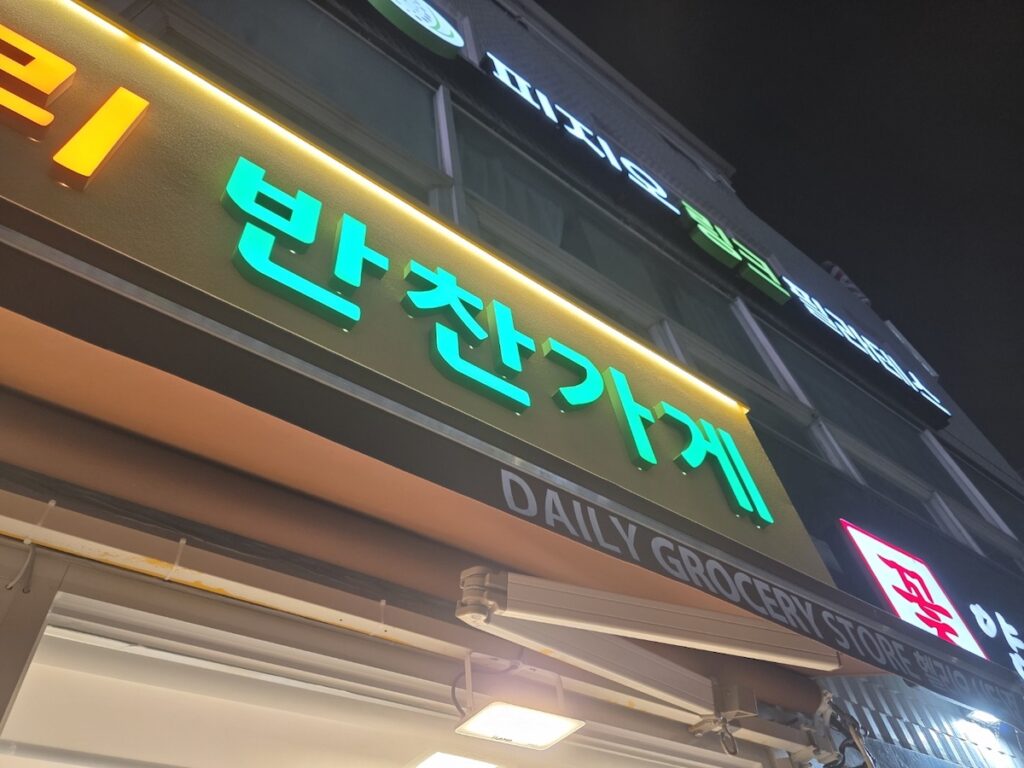 An angled nighttime cinematic shot of a neighborhood Banchan-gage in Seoul. The glowing green LED sign clearly reads '반찬가게' (Side Dish Shop), with 'DAILY GROCERY STORE' in white text below it, representing the local source for an authentic Korean weight loss diet banchan selection.