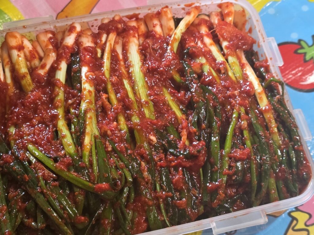 The Most Delicious Kimchi showing a container full of freshly seasoned Korean green onion kimchi (Pa-kimchi), highlighting the vibrant red chili flakes and "Two-Tone" green and white stalks.