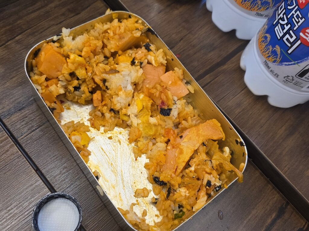 A close-up of a 1970s-style nickel-silver lunchbox (Dosirak) filled with white rice, fried egg, pink sausage, and spicy sauce, served as a nostalgic meal after Korean BBQ.