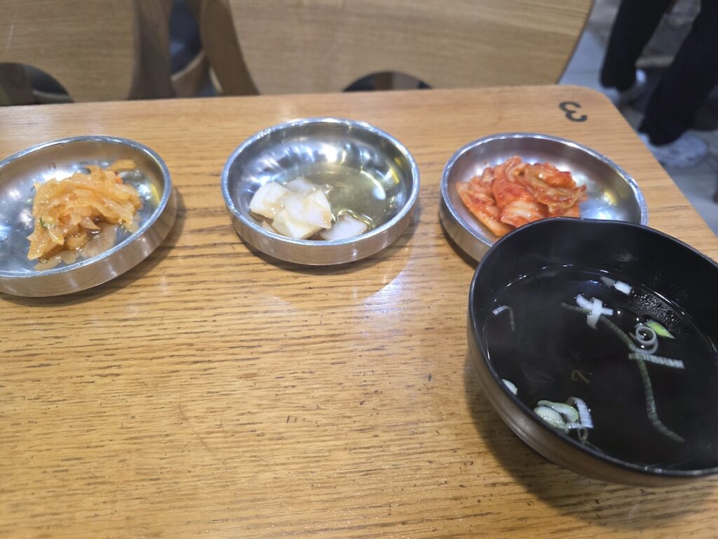 A metal tray holding three tiny, uninspiring metal bowls of basic side dishes (radish, pickled onion, commercially produced kimchi) and a bowl of plain soup, contrasting with generous Korean meal traditions.