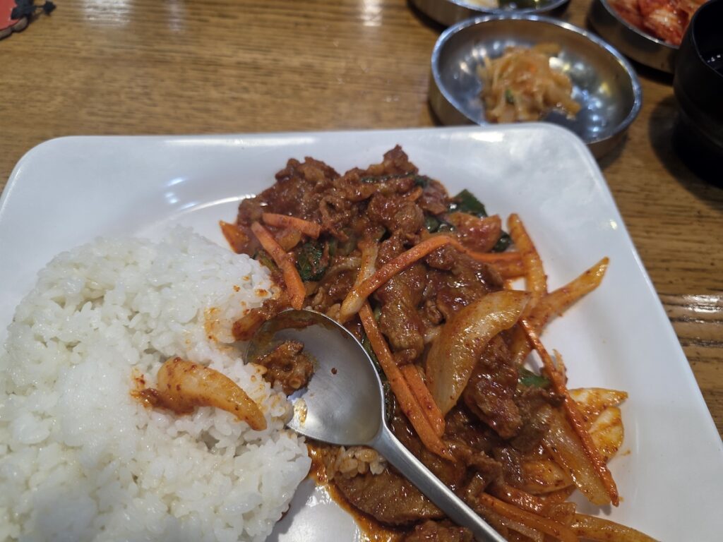 A close-up view of a mediocre serving of Spicy Pork (Jeyuk-bokkeum) with rice on a white plate at a Korean bus terminal restaurant, served with meager side dishes, showcasing a common tourist trap in Korea.