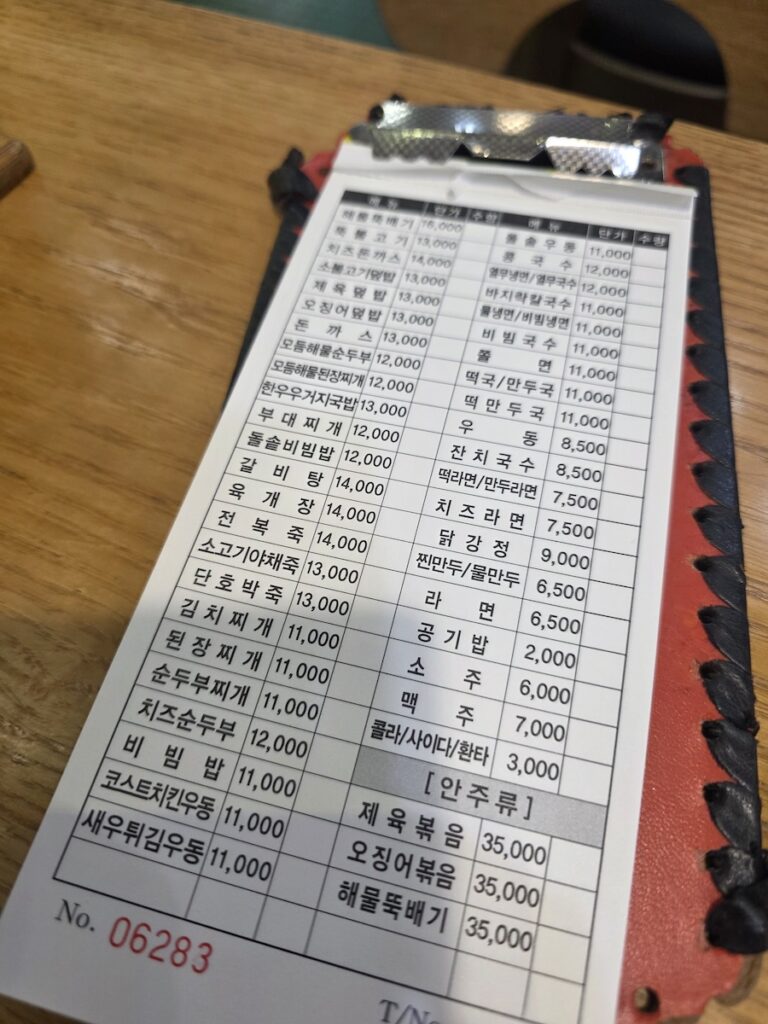 A close-up of an old, cluttered paper order sheet on a wooden table, with a hand holding a pen over high-priced standard items like 13,000 KRW Jeyuk-bokkeum at a South Korean transit hub restaurant.