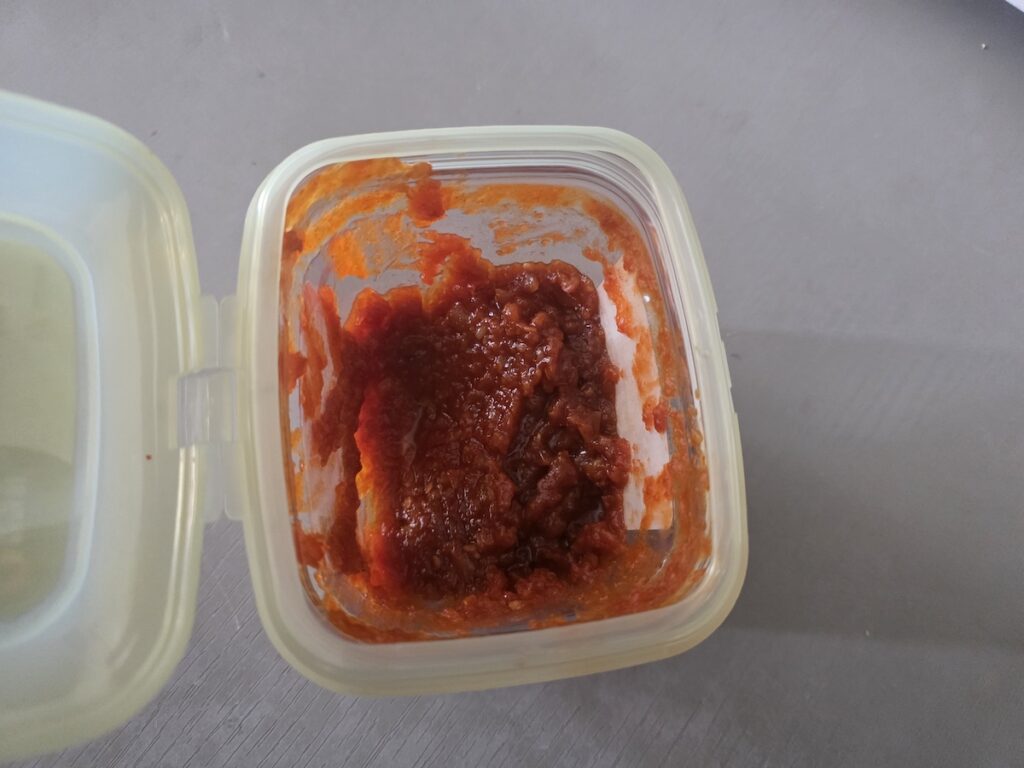An open plastic container on a gray table filled with savory, reddish-brown Korean Ssamjang sauce, a fermented plant-based dip for vegetables.