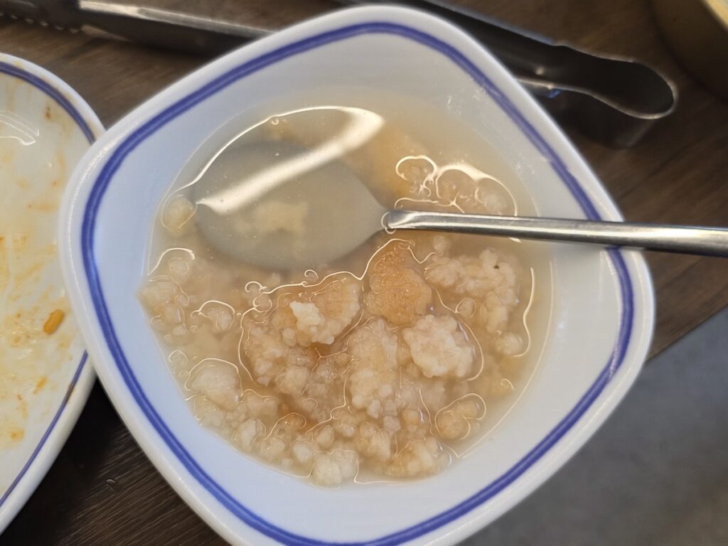 A white bowl of warm scorched rice soup (Nurungji) with a spoon, showing the soft and comforting texture of a traditional meal after Korean BBQ.