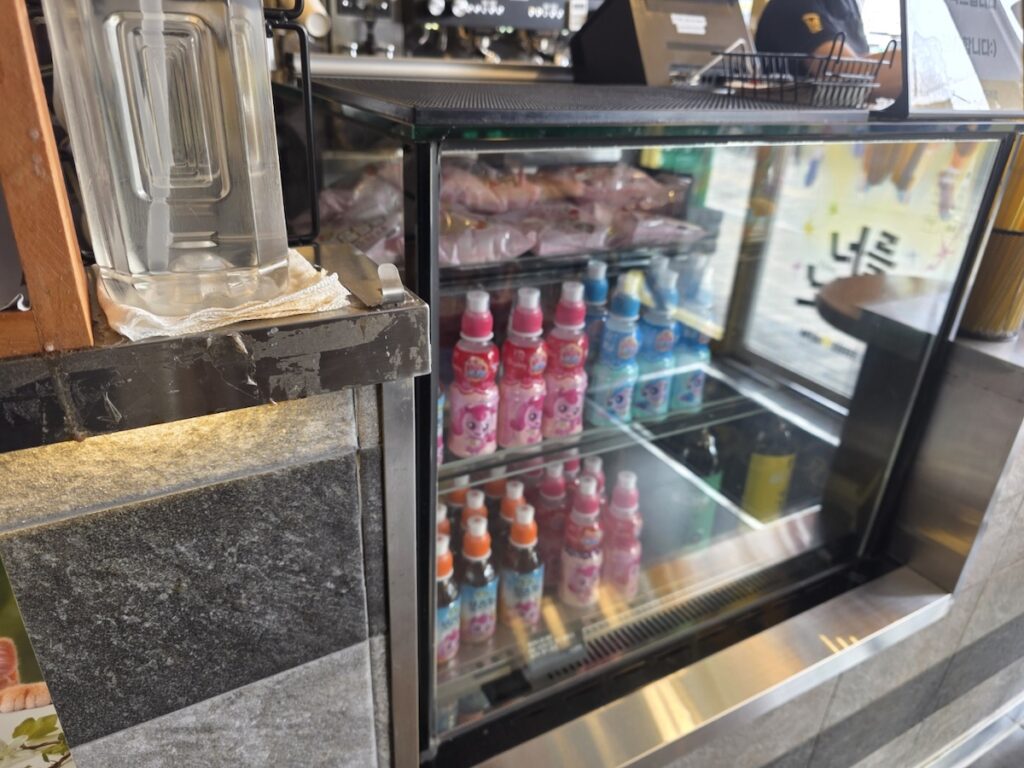 A refrigerated showcase at a Mega Coffee Korea branch, displaying colorful bottled character drinks for kids, part of the extensive Mega Coffee Korea Dessert Menu.