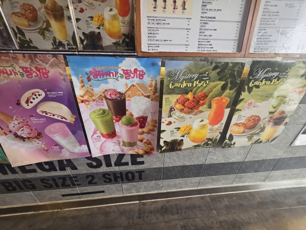 Multiple colorful seasonal drink posters, including strawberry and tea variations, displayed on the wall of a Mega Coffee Korea shop, showcasing the vibrant Mega Coffee Korea Dessert Menu.