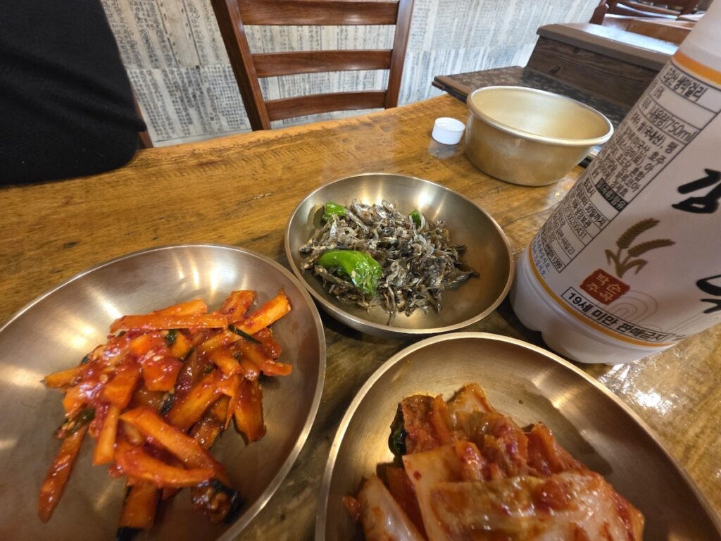 A side view of seasoned anchovies, spicy radish, and kimchi in brass plates next to a local Makgeolli bottle, illustrating the pairing section of a Regional Makgeolli Guide.