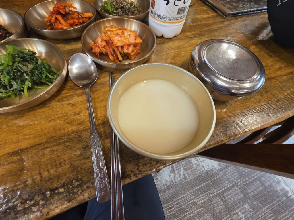 A brass bowl filled with milky white Makgeolli, positioned alongside traditional side dishes and a silver rice bowl, as explained in this Regional Makgeolli Guide.