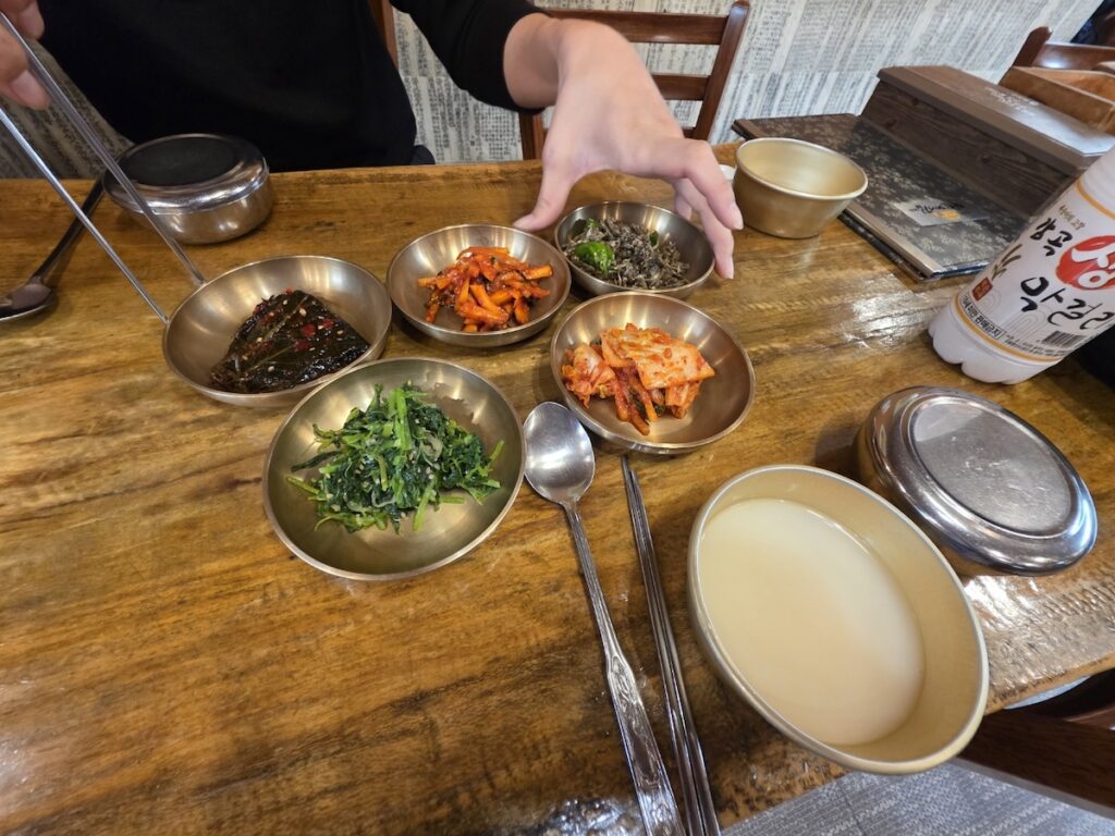 An overhead shot of a full Korean meal setting with five types of banchan in brass bowls and a bowl of Makgeolli, showcasing the experiential side of a Regional Makgeolli Guide.