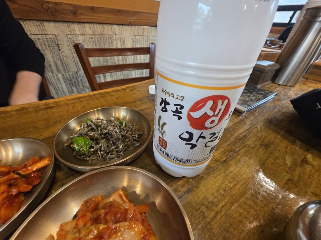 A close-up of a white bottle of Gamgok Saeng Makgeolli on a wooden table, part of a Regional Makgeolli Guide, surrounded by brass bowls of Korean side dishes like kimchi and anchovies.