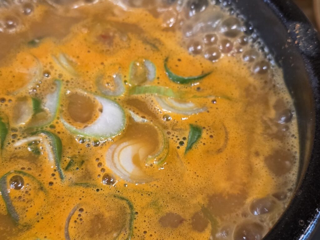 A detailed view of bubbling orange broth with sliced green onions in a what is why how guide explained about the sensory experience of eating Korean fermented stew.